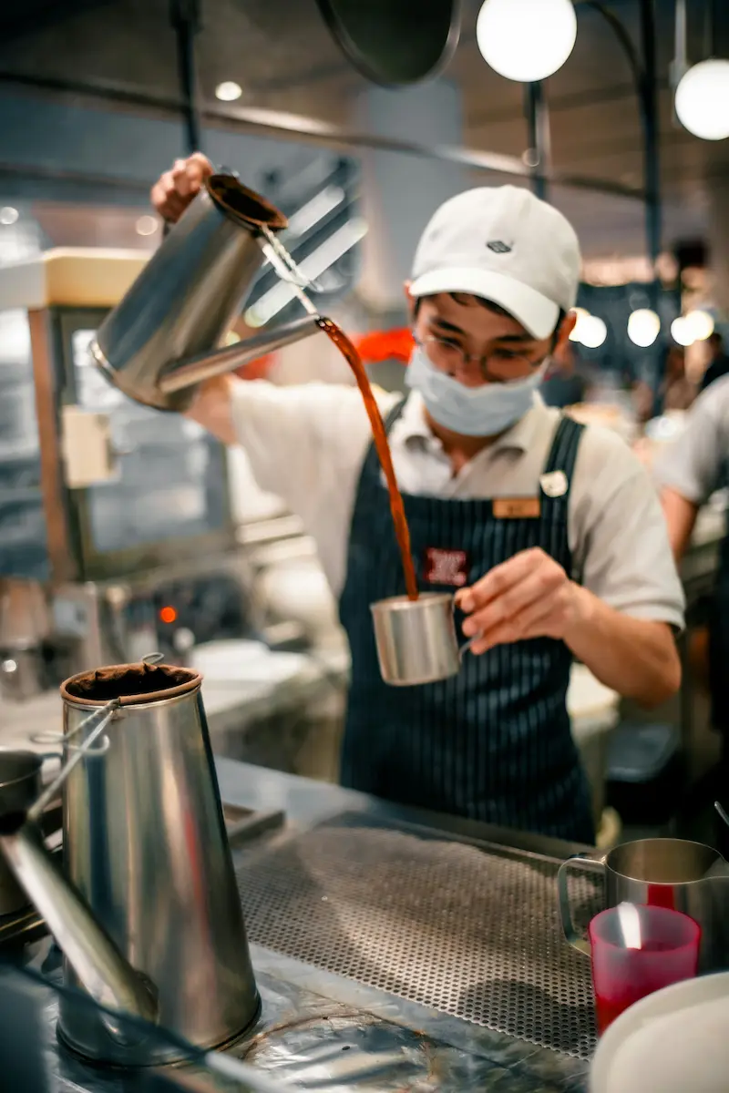 A kopitiam drinks stall worker pouring a hot drink from a metal pot into a metal cup
